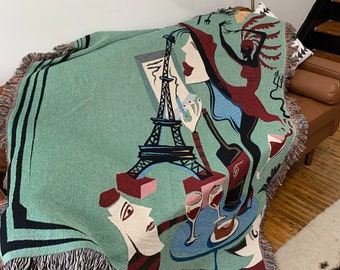 May include: A woven throw blanket with a light green background and a colorful design featuring the Eiffel Tower, wine bottles, glasses, and figures. The blanket has a fringed edge and is draped over a brown leather couch.