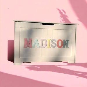 May include: White wooden toy chest with a hinged lid and a cut-out handle. The front of the chest has the name "MADISON" spelled out in colourful letters.
