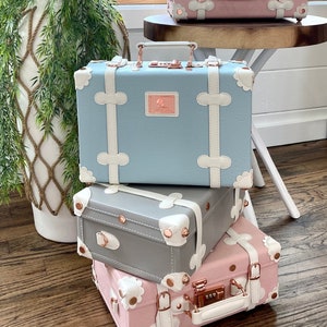 May include: Three vintage-style suitcases stacked on top of each other. The top suitcase is light blue with white trim and a rose gold lock. The middle suitcase is gray with white trim and a rose gold lock. The bottom suitcase is pink with white trim and a rose gold lock. Each suitcase has a white flower design on the front.