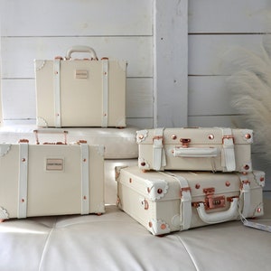May include: Four vintage-style suitcases in various sizes, all white with rose gold accents and hardware. The suitcases are arranged in a pyramid shape on a white couch.