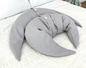 May include: Two crescent moon-shaped pillows in a solid light gray color. Each pillow features a small rectangular fabric tag sewn onto the center of the outer curve. The pillows are made of a textured fabric and have a curved, pointed design.