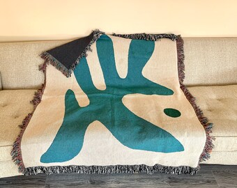 May include: A woven throw blanket with a teal abstract design on a cream background. The blanket has fringe around the edges and is draped over a couch.