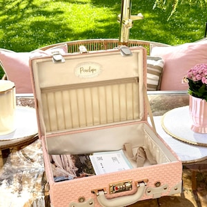 Puede incluir: Un maletín de aseo de color rosa claro con textura, asa superior y cerradura de combinación. El interior cuenta con un forro color crema, un bolsillo de malla con cremallera y una placa central con el nombre Penelope en cursiva.