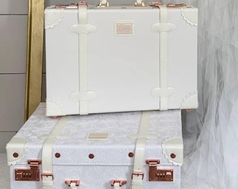 May include: Two white vintage-style suitcases with rose gold hardware are stacked on a white upholstered bench. A gold-framed display holds a white corset and a flowing white fabric. Floral arrangements with pink and green accents adorn the scene.