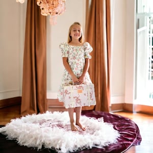 May include: A young girl in a white floral dress stands on a white and burgundy rug, holding a vintage-style suitcase with pink floral accents.
