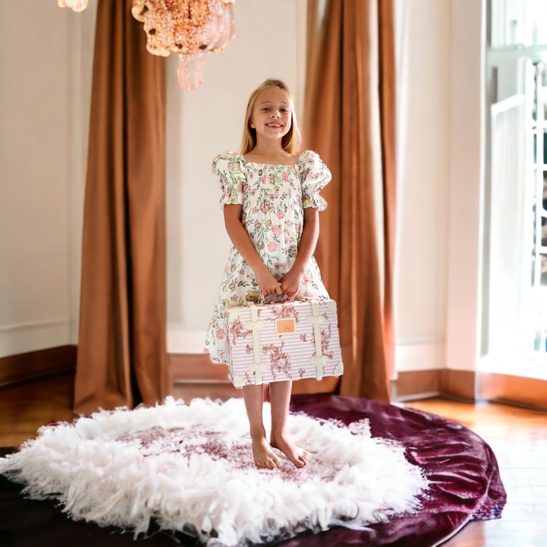 May include: A young girl in a white floral dress stands on a white and burgundy rug, holding a vintage-style suitcase with pink floral accents.