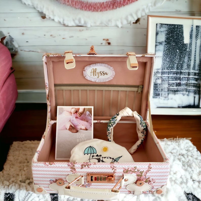 May include: A pink and white striped vintage-style suitcase with a gold lock and handle. The suitcase is open and contains a headband, a small white bag with a bear and umbrella design, and a photo of a baby girl wearing a pink tutu. The suitcase has a personalized name tag that says "Alyssa".