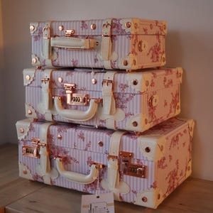 May include: A set of three vintage-style suitcases with pink and white floral patterns and gold accents. The suitcases are stacked on top of each other.
