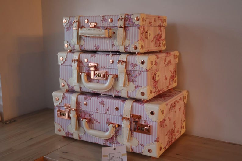 May include: A set of three vintage-style suitcases with pink and white floral patterns and gold accents. The suitcases are stacked on top of each other.