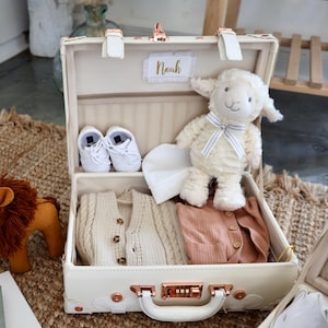May include: A white vintage suitcase with gold accents, personalised with the name "Noah". The suitcase is open and filled with baby items, including a white knitted sweater, a brown onesie, a white hat, and a white stuffed lamb.