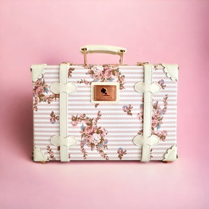 May include: A white vintage-style suitcase with a floral and striped pattern. The suitcase has a gold-colored handle and metal accents. The suitcase has a small gold-colored tag with the text "Since 1978" on it.