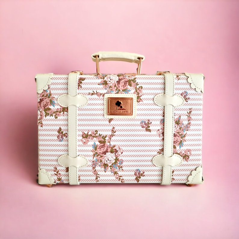 May include: A white vintage-style suitcase with a floral and striped pattern. The suitcase has a gold-colored handle and metal accents. The suitcase has a small gold-colored tag with the text "Since 1978" on it.