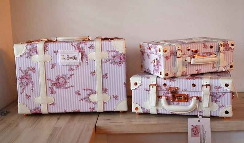 May include: Three vintage-style suitcases with a pink and white floral pattern and gold accents. The largest suitcase has a label that reads "The Smiths".