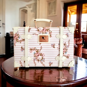 May include: A white vintage-style suitcase with a pink and white floral pattern and pink stripes. The suitcase has a gold handle and gold accents. The suitcase has a small gold plaque with the text "Since 1979".