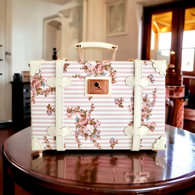 May include: A white vintage-style suitcase with a pink and white floral pattern and pink stripes. The suitcase has a gold handle and gold accents. The suitcase has a small gold plaque with the text "Since 1979".