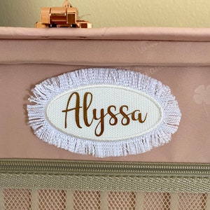 May include: A white oval luggage tag with a frayed white border. The tag has the name "Alyssa" written in gold lettering.
