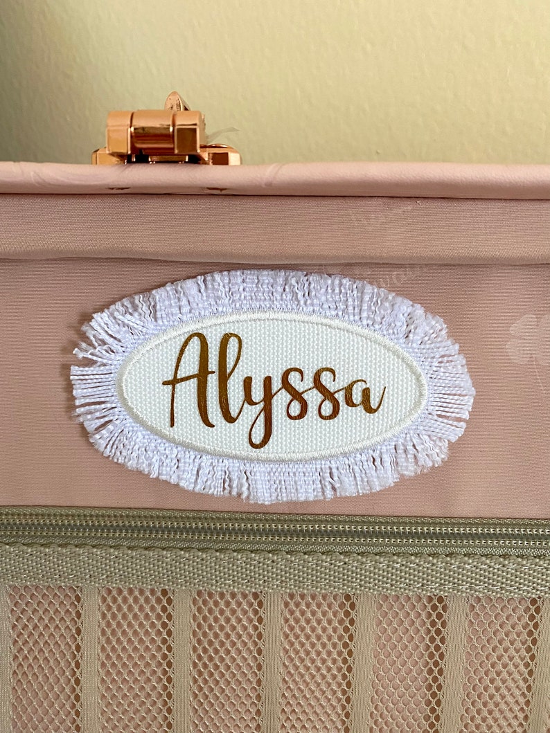 May include: A white oval luggage tag with a frayed white border. The tag has the name "Alyssa" written in gold lettering.