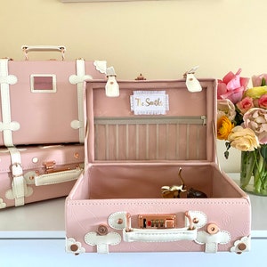 May include: Three pink vintage-style suitcases with white trim and floral accents. The top suitcase is open and has a white label with the text "The Smiths".