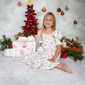 May include: A young girl wearing a white dress with pink floral print sits in front of a Christmas tree decorated with red ornaments. She is smiling and looking at the camera. A pink and white striped suitcase with floral accents is in the foreground.