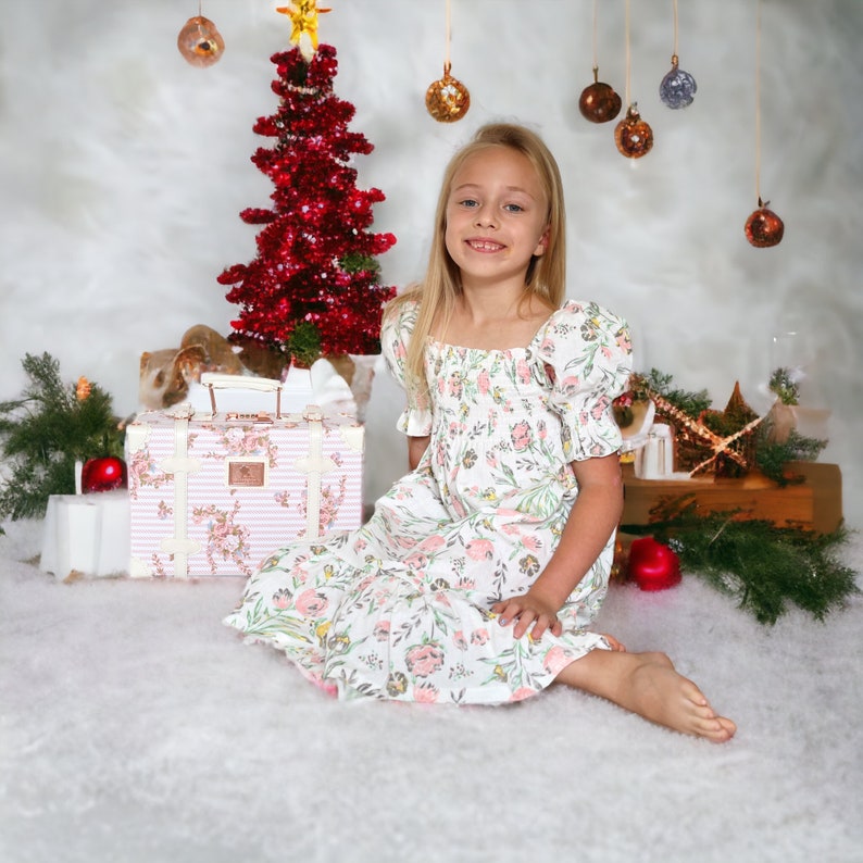 May include: A young girl wearing a white dress with pink floral print sits in front of a Christmas tree decorated with red ornaments. She is smiling and looking at the camera. A pink and white striped suitcase with floral accents is in the foreground.