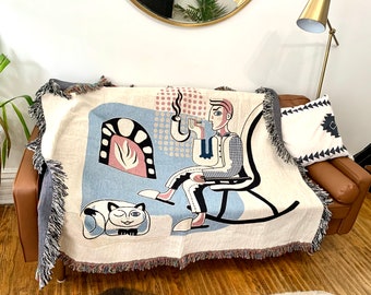 May include: A woven throw blanket with a colorful geometric design featuring a person sitting in a rocking chair, a cat, and a fireplace. The blanket has a fringe border.