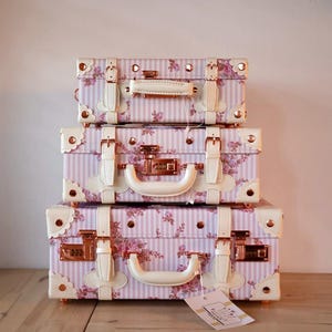 May include: Three vintage-style suitcases stacked on top of each other. The suitcases are pink and white with a floral pattern and have gold accents. The suitcases have white leather handles and gold locks.