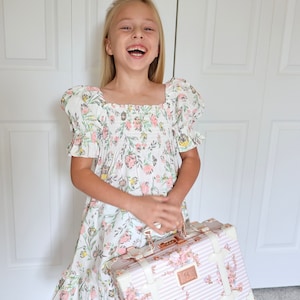 May include: A young girl wearing a white dress with pink and green floral print and short puffed sleeves is holding a pink and white floral patterned suitcase with gold accents.