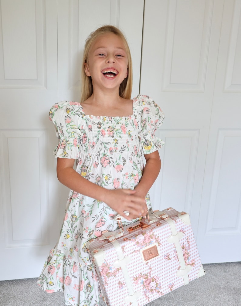 May include: A young girl wearing a white dress with pink and green floral print and short puffed sleeves is holding a pink and white floral patterned suitcase with gold accents.