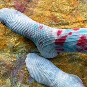 Könnte beinhalten: Ein Paar hellblaue gerippte Socken mit Batikmustern in Rot- und Türkistönen. Die Socken werden vor einem strukturierten, goldfarbenen Hintergrund präsentiert. Die Socken sind aus einem weichen, bequemen Material gefertigt.