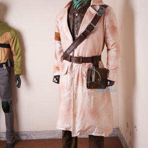 May include: A long, light-colored coat with a weathered finish worn over a dark brown vest and dark trousers. The outfit includes a dark brown leather shoulder strap, a matching belt with a pouch, and a green neck scarf on a dark mannequin.