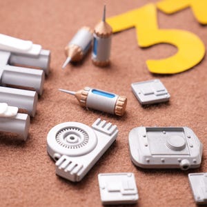 May include: An assortment of gray and white miniature machine parts, including cylinders, rectangular blocks, and a gear-shaped component, are scattered on a brown surface. Two yellow numbers, "55" and a black and yellow "33", are also present.