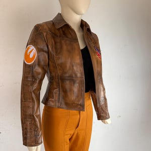 May include: A brown leather jacket with a collared neckline and a fitted silhouette. The jacket features a patch on the left sleeve with an orange and white design, and a patch on the right side. The jacket is paired with orange trousers.