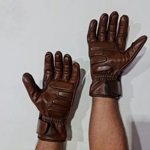 Könnte beinhalten: Ein Paar braune Lederhandschuhe mit einem strukturierten Design auf dem Handrücken. Die Handschuhe haben einen Handgelenksriemen und werden an den Händen einer Person gezeigt.