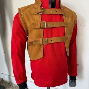 May include: A brown suede vest with a high collar and multiple buckled straps. The vest is worn over a red long-sleeved shirt.