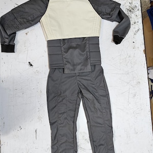 Puede incluir: Un traje protector gris y crema, extendido sobre una superficie blanca. El traje incluye una chaqueta de cuello alto con un panel de pecho color crema, mangas grises y pantalones grises a juego. El traje parece estar hecho de un material duradero, posiblemente resistente al agua.