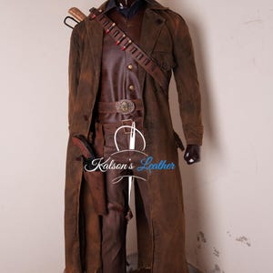 May include: A brown distressed leather duster coat worn over a dark vest and pinstriped trousers. The outfit includes a leather bandolier with red-tipped cartridges, a belt with a decorative buckle, and a holster containing a prop revolver.