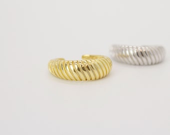 Könnte beinhalten: Zwei offene Ringe mit einer dicken, abgerundeten und gerippten Textur. Ein Ring ist in einem hellen Goldton gehalten, während der andere in einem polierten Silberton ausgeführt ist. Beide Ringe weisen ein durchgehendes diagonales Rillenmuster auf.