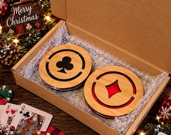 May include: Two circular wooden coasters inside a cardboard box, one featuring a black club symbol and the other a red diamond symbol. The coasters are nestled in bubble wrap, surrounded by playing cards and Christmas decorations.