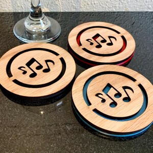 May include: Three wooden coasters with a musical note design. Each coaster has a light brown wood grain top with a black border and a colored accent ring: red, blue, and black. The coasters are round and sit on a dark surface.