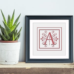 May include: A framed print with a red letter A in a decorative frame with a white background. The frame is black with a white mat.