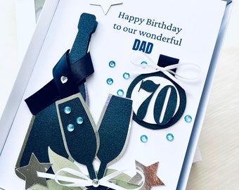 May include: A white card with a blue and silver design. The card says "Happy Birthday to our wonderful Dad" and "70". There are two champagne glasses and a bottle of champagne on the card.