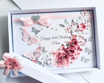 May include: A handmade card with a pink and white floral design, butterflies, and glitter. The card is in a white box and says "Happy 80th Birthday Mum".