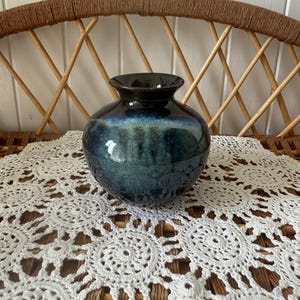 May include: A round, glazed ceramic vase with a dark blue and green colour gradient. The vase has a wide body and a narrow neck. It sits on a white, crocheted doily, with a woven wicker chair in the background.