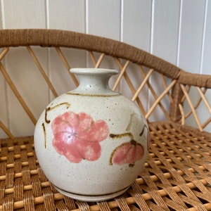 May include: A small, round ceramic vase with a speckled white glaze and brown accents. The vase is decorated with pink floral designs.