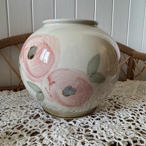May include: A white ceramic vase with a round body and a wide opening. The vase is decorated with pink and green floral designs.
