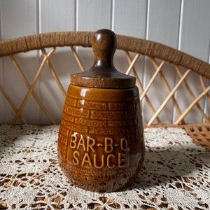 May include: A brown ceramic jar with a wooden lid. The jar has a brick pattern and the text "BAR-B-Q SAUCE" is printed on the front.