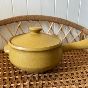 May include: A yellow ceramic casserole dish with a lid and a handle. The dish is sitting on a woven wicker surface.