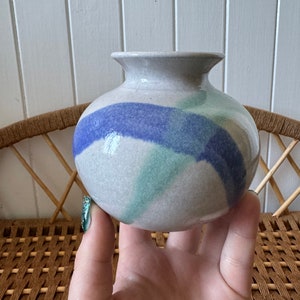 May include: A small, round ceramic vase with a light grey glaze and abstract blue and green brushstrokes.