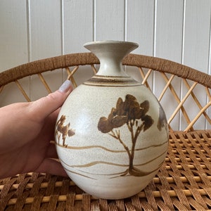May include: A white ceramic vase with a brown glaze and a hand-painted design of trees and a landscape. The vase has a round body and a narrow neck.