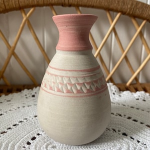 May include: A small, white ceramic vase with a pink rim and a pink geometric design around the middle. The vase is sitting on a white crocheted doily.